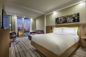 A hotel room with a large bed, a desk with a chair, a desk lamp, a telephone, and a cityscape view through the window. - Hampton by Hilton Istanbul Atakoy (Istanbul)