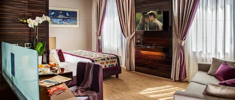 A hotel room with a bed, a sofa, a TV, a desk with a computer, and a window with curtains.