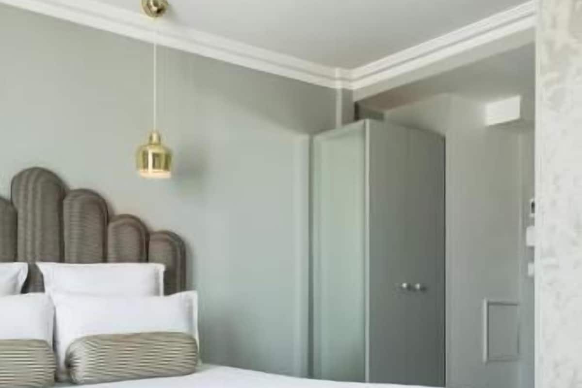 A neatly made bed with a grey skirt, a hanging light fixture, and a wardrobe in a room with a light-colored wall.