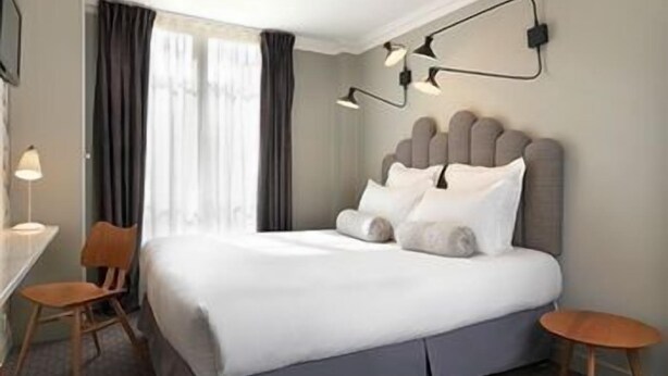 A neatly arranged hotel room with a bed, bedside table, chair, and lamp. - Paradise Hotel (Paris)