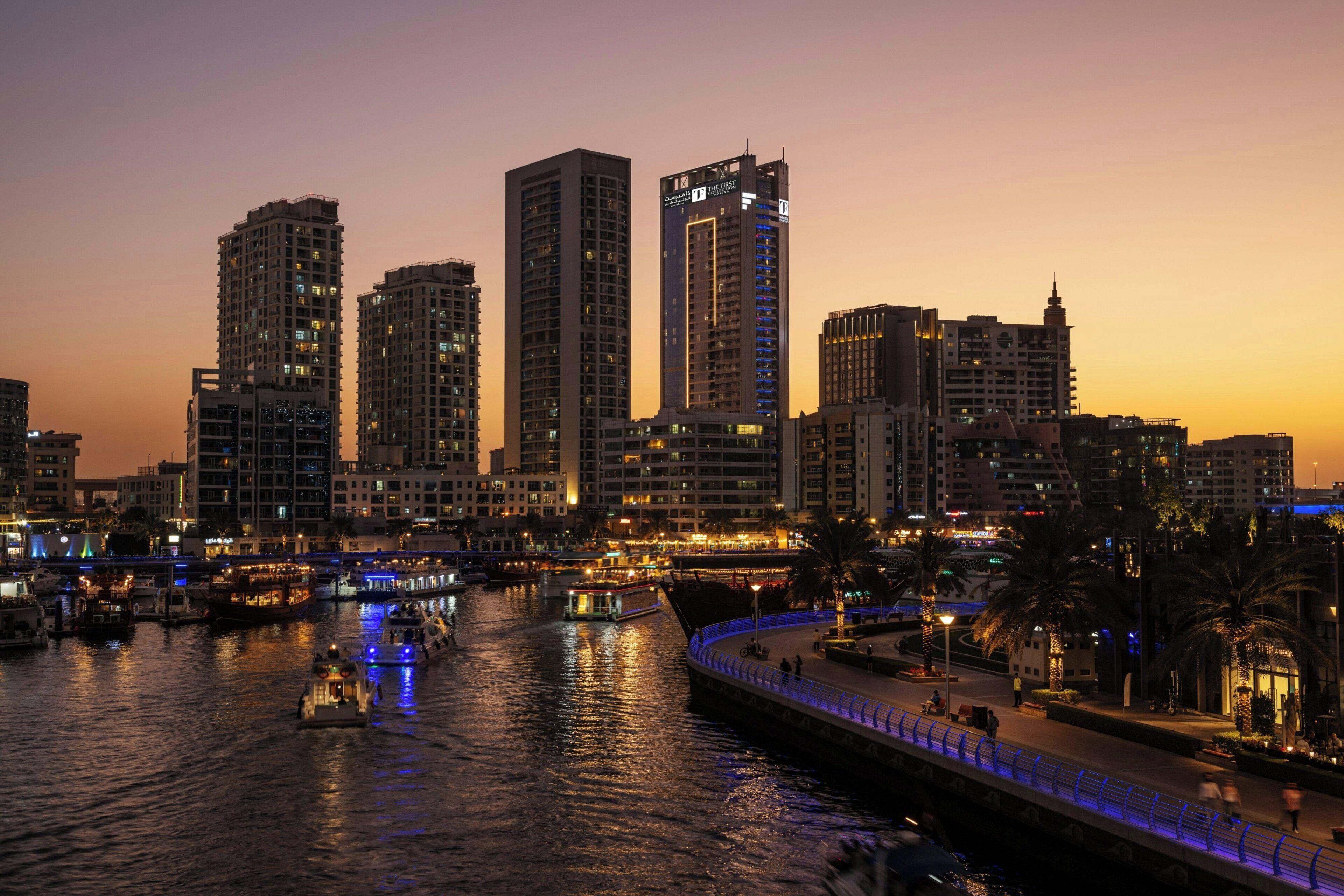 A cityscape at dusk with high-rise buildings, a waterfront promenade, and boats.