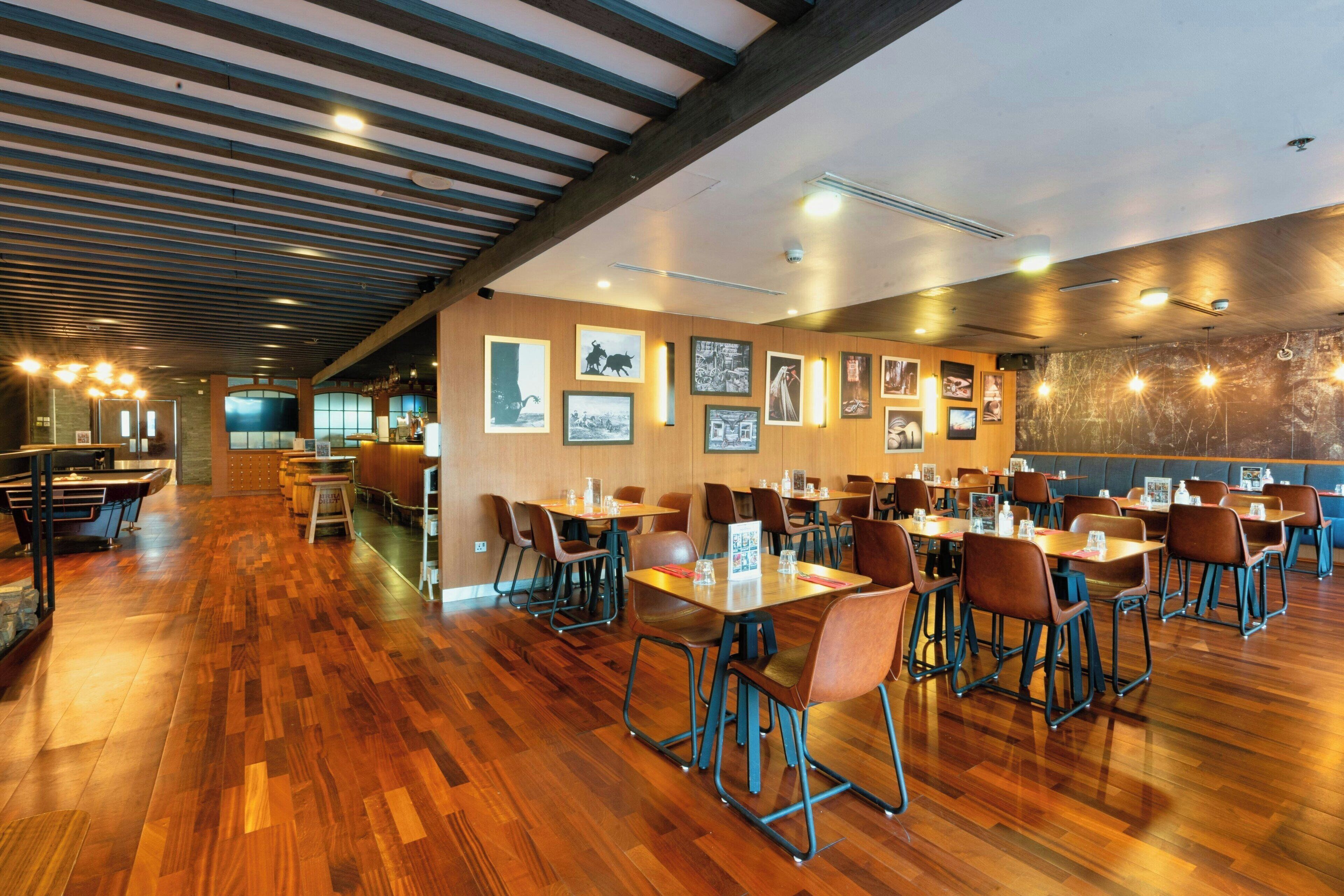 A spacious restaurant with wooden flooring, multiple tables and chairs, and framed pictures on the walls.