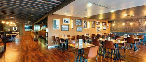 A spacious restaurant with wooden flooring, multiple tables and chairs, and framed pictures on the walls.