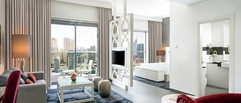 A modern hotel room with a city view, a flat-screen TV, a sofa, a red armchair, and a dining area with a coffee table.
