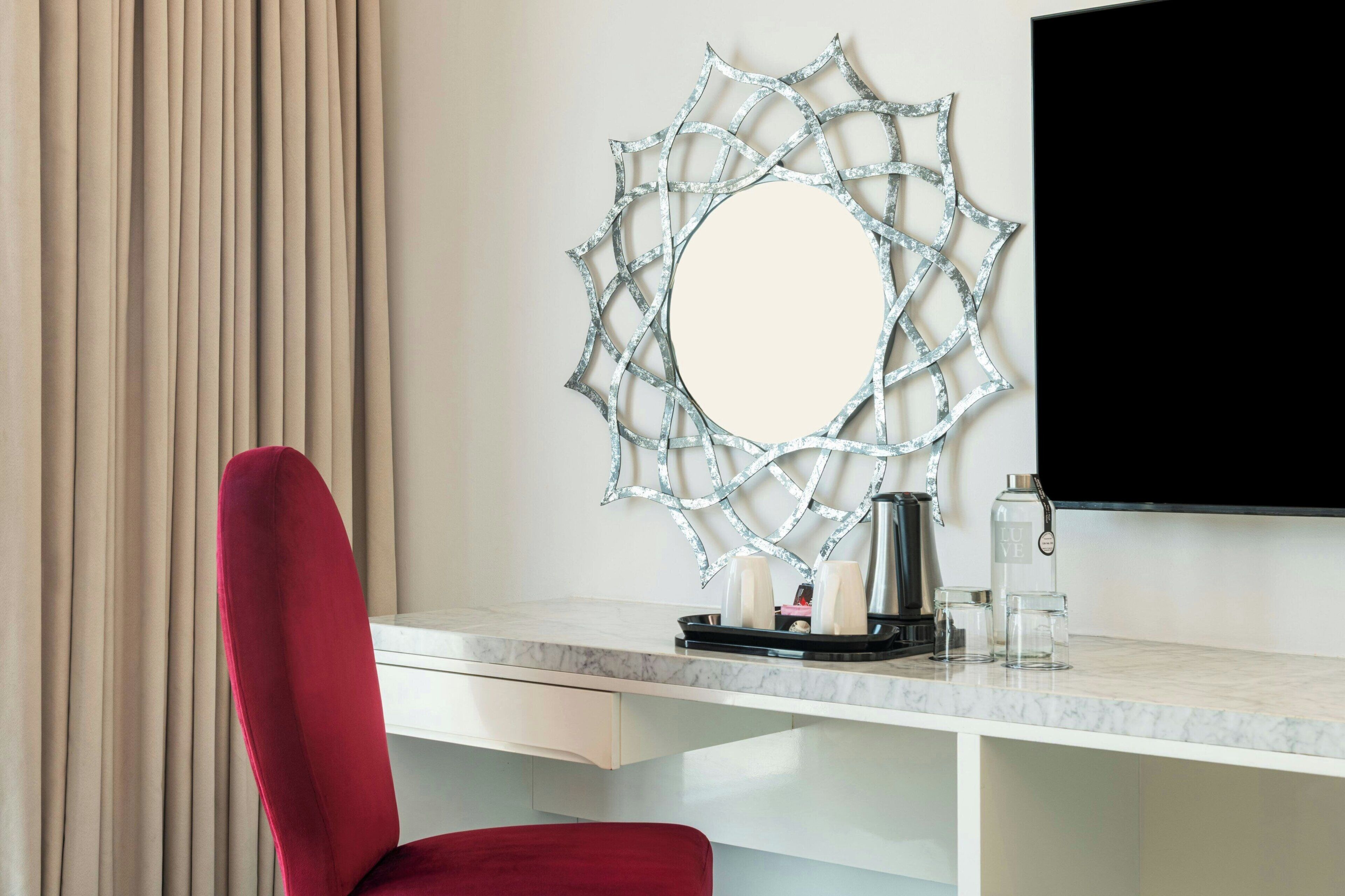 Habitación de hotel con una silla roja, un escritorio blanco, un espejo grande con diseño de telaraña, un televisor de pantalla plana y una cafetera.