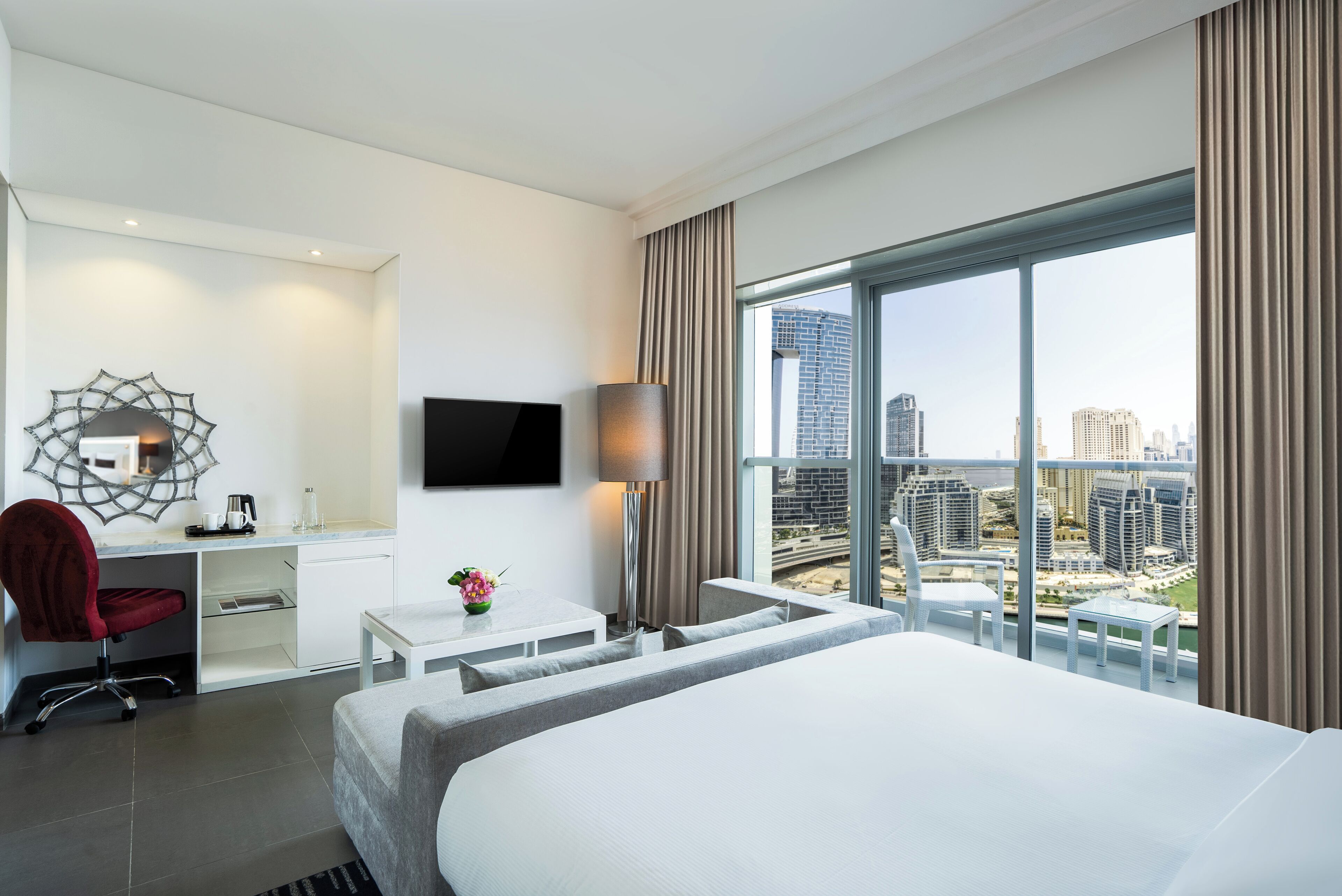 A modern hotel room with a large bed, a desk with a chair, a TV, and a view of the cityscape.