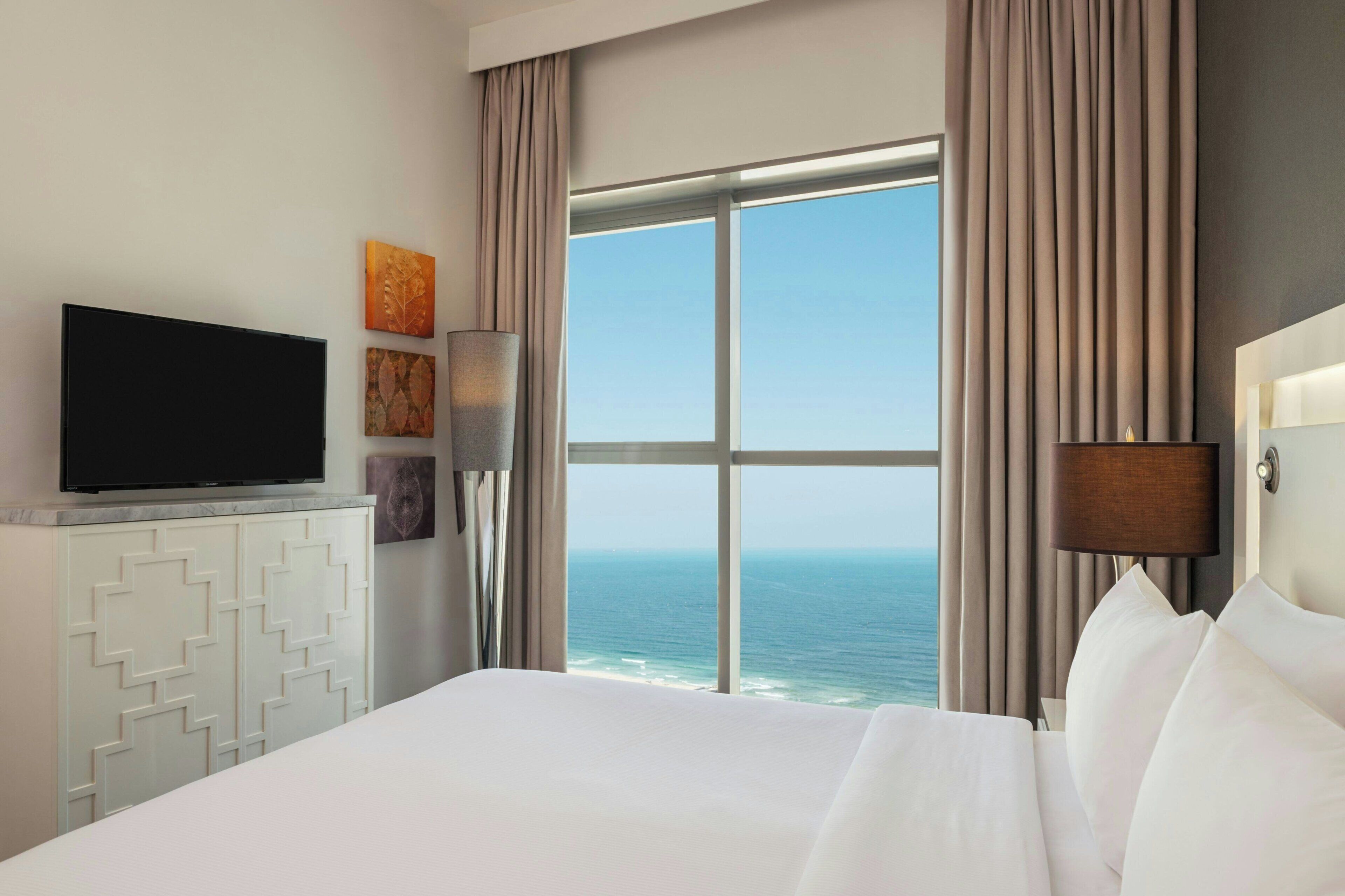 Habitación de hotel con una cama grande, un televisor sobre un mueble blanco, vista al océano y una ventana con cortinas.