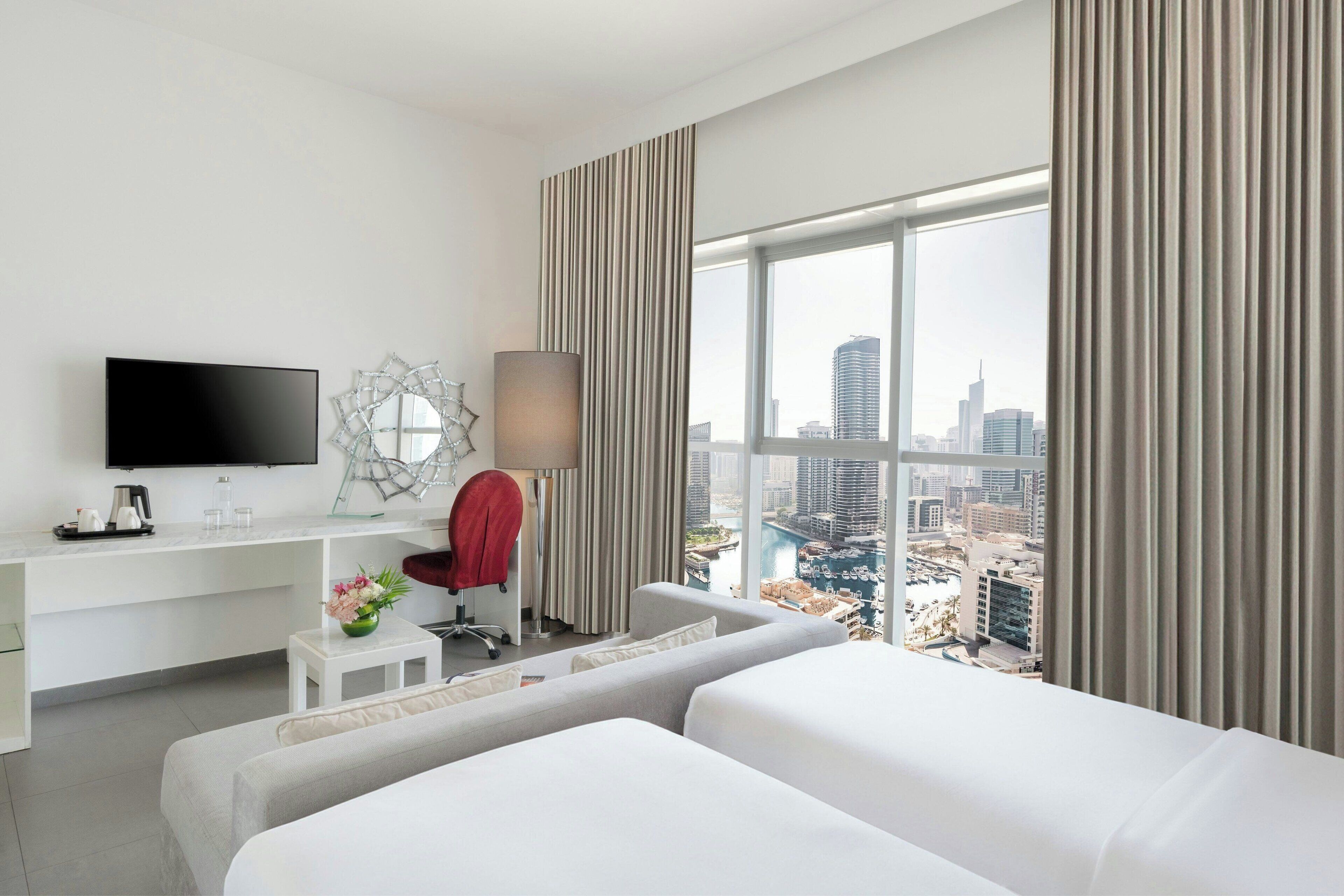 Une chambre d’hôtel moderne dotée d’une grande fenêtre, d’un téléviseur à écran plat, d’un bureau avec une lampe et d’un fauteuil rouge.