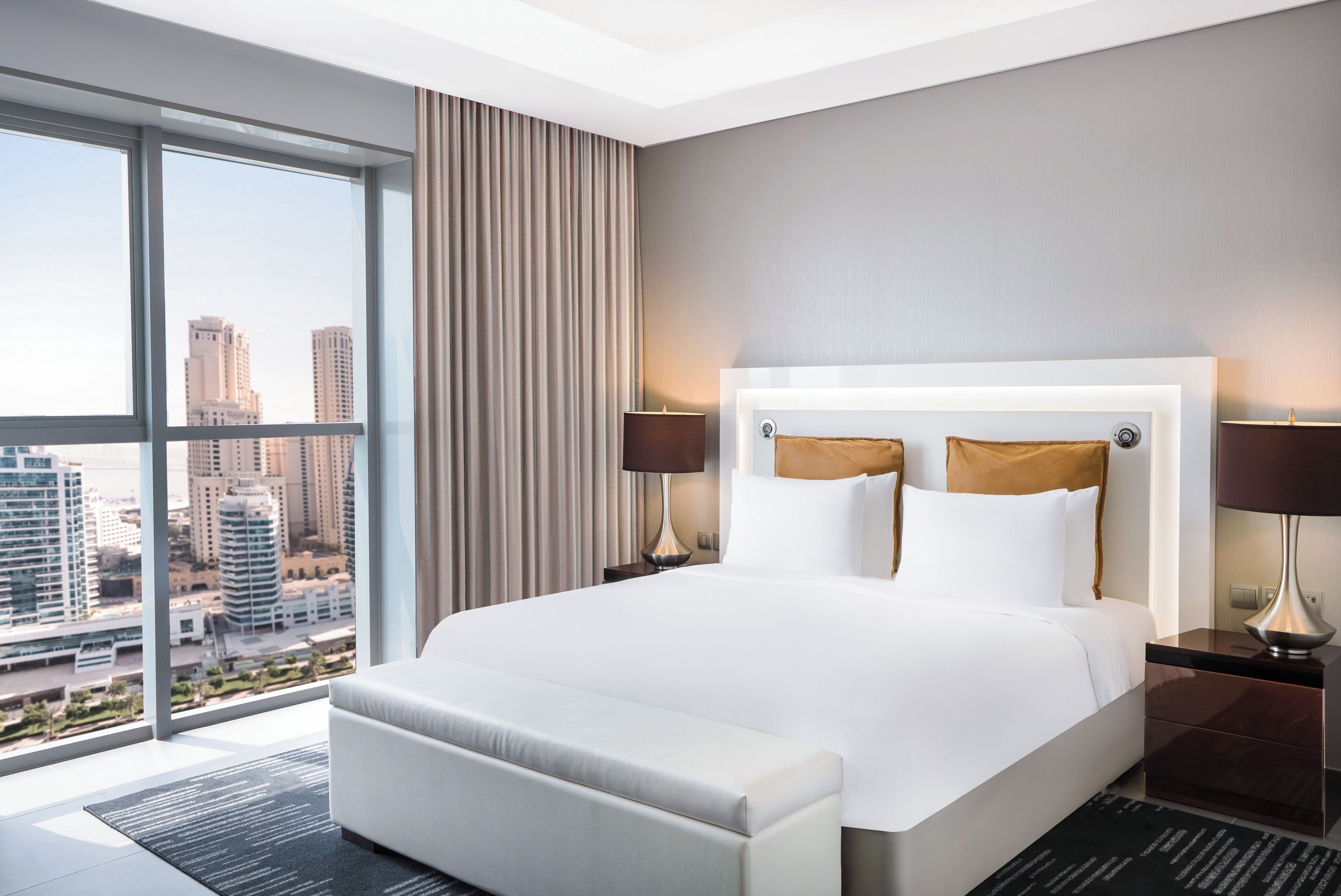 A modern hotel room with a large bed, two bedside lamps, a nightstand, and a view of the cityscape through floor-to-ceiling windows.