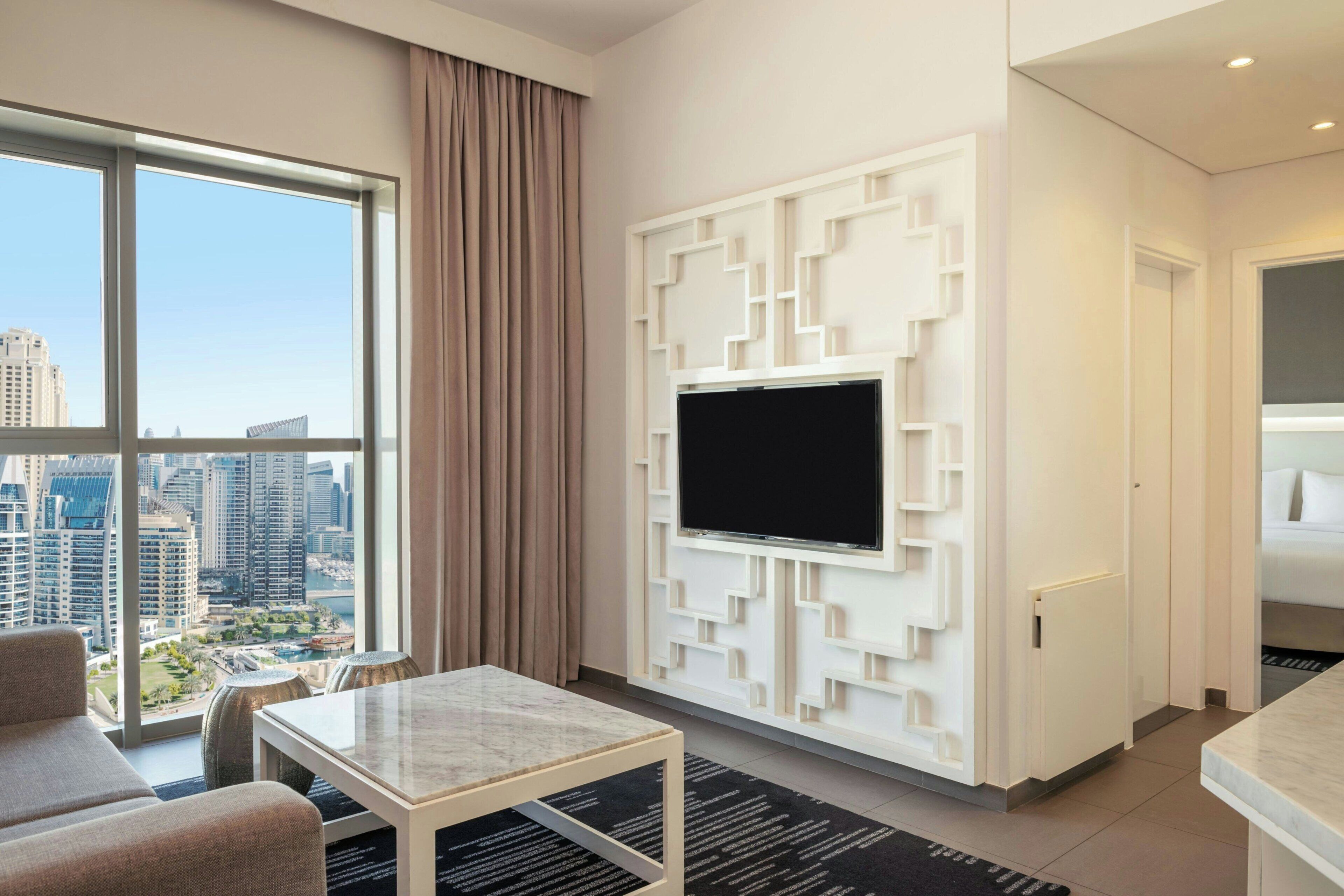 A modern hotel room with a large window, a flat-screen TV mounted on a geometric wall, a sofa, and a small table.