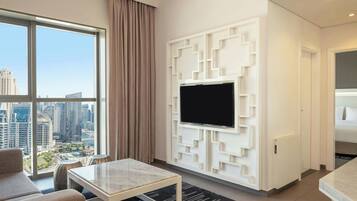 A modern hotel room with a large window, a flat-screen TV mounted on a geometric wall, a sofa, and a small table.