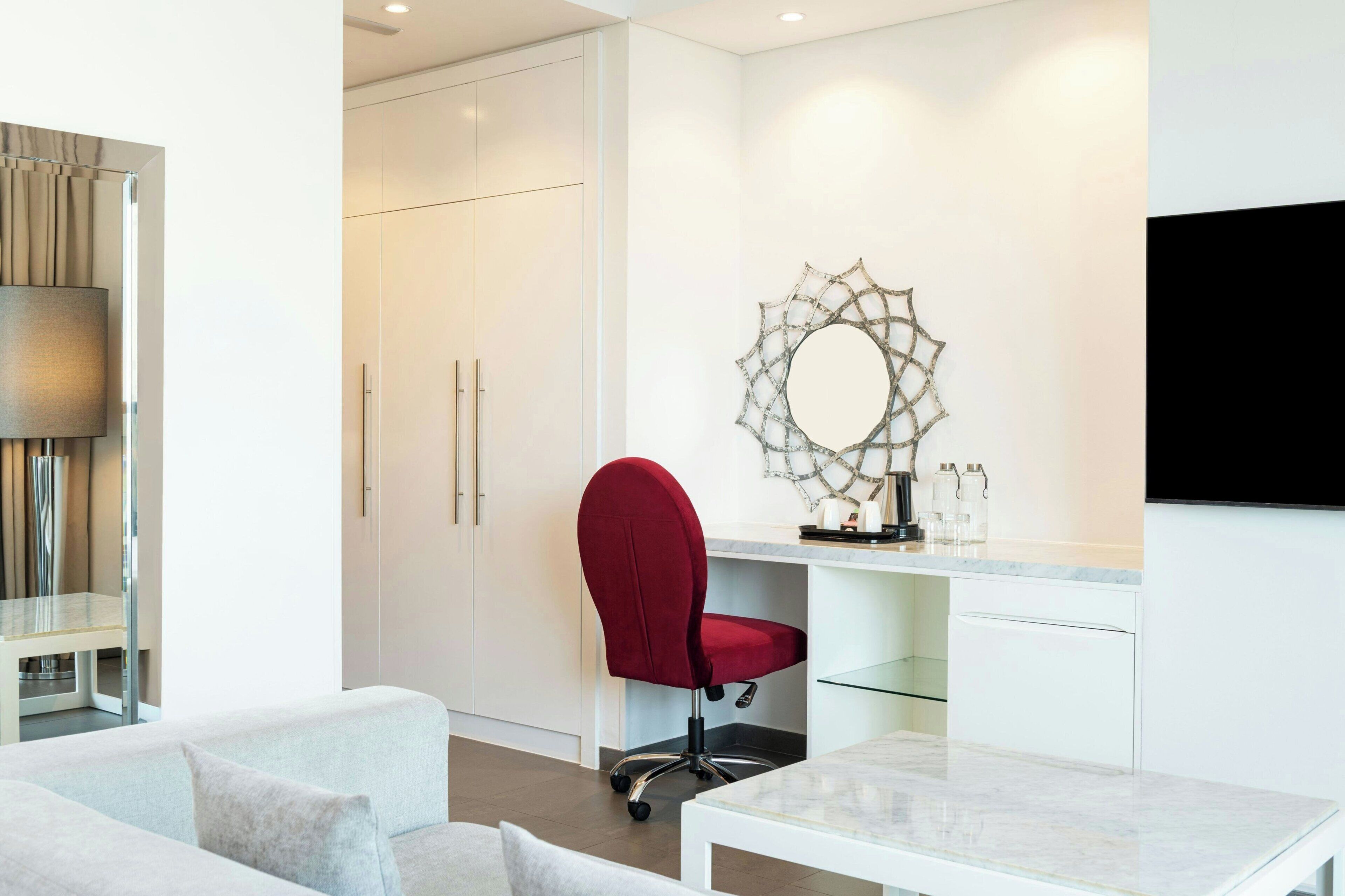 A modern room with a red office chair, a glass table, a white desk, and a wall-mounted TV.