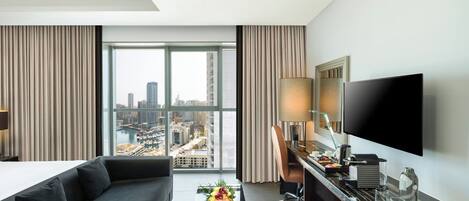 A modern hotel room with a large window, a sofa, a desk with a flat-screen TV, and a coffee maker.