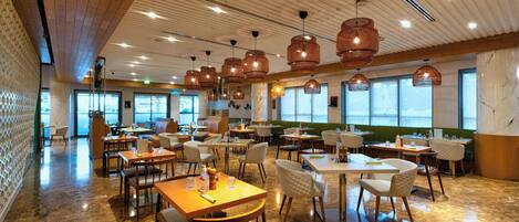 Ein geräumiges Restaurant mit hohen Decken, Holzbalken und großen Fenstern. Tische und Stühle sind ordentlich angeordnet, und hängende Pendelleuchten sind sichtbar.