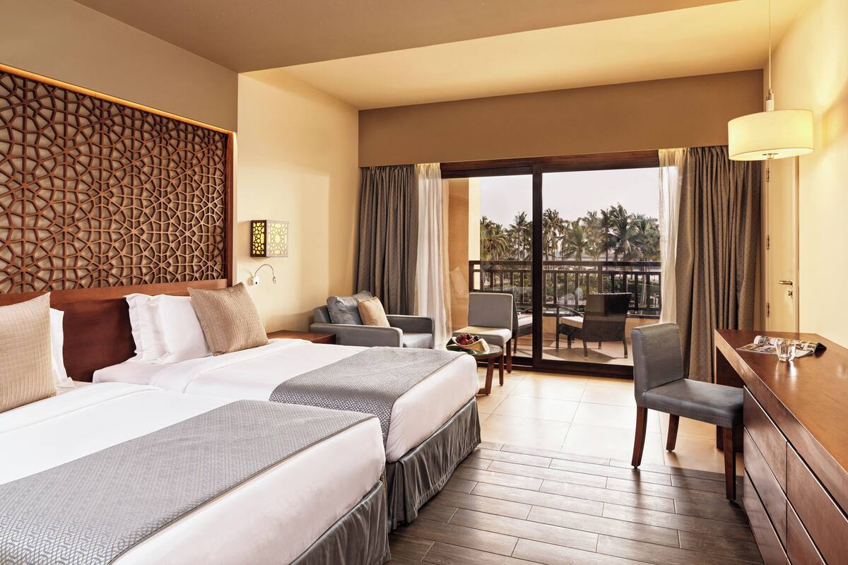 A hotel room with a large bed, a balcony with a view of palm trees, and a desk with a lamp.