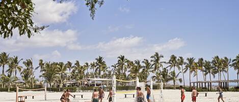 Partido de voleibol de playa con jugadores en traje de baño, palmeras y una red de voleibol en una playa arenosa.