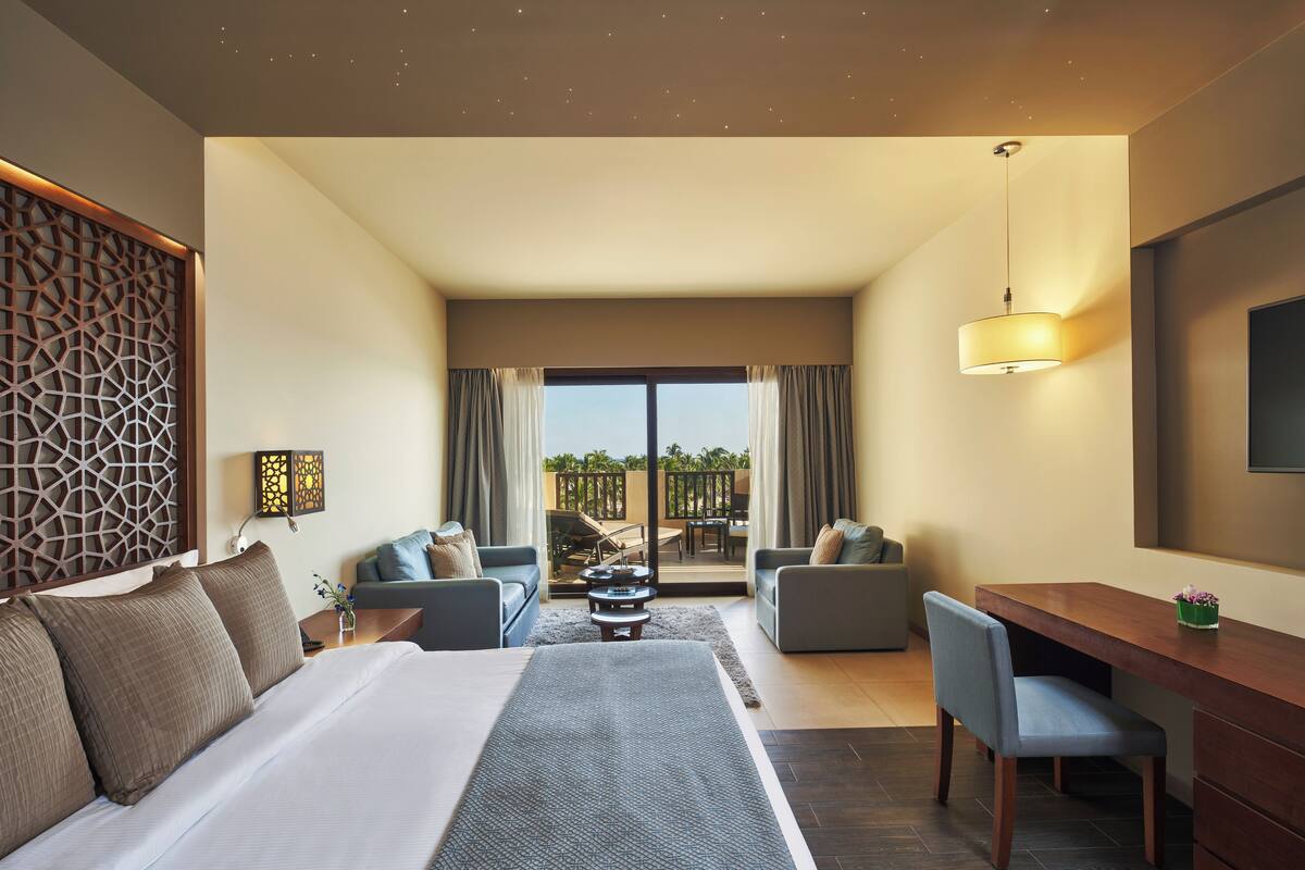 A hotel room with a large bed, two sofas, a desk, and a balcony with outdoor furniture.