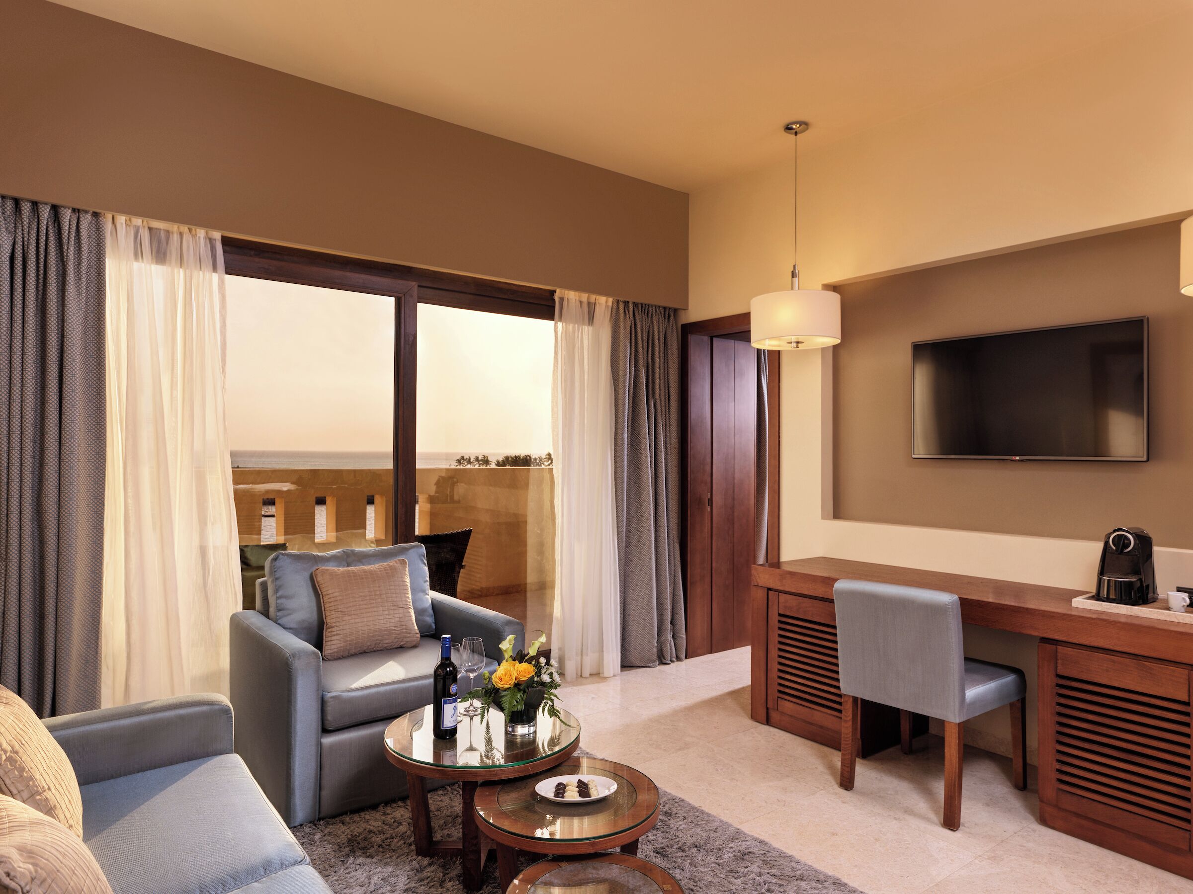 Une chambre d’hôtel avec un canapé, une table à manger, une télévision et un balcon avec des rideaux.