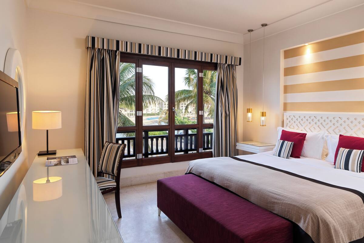 A hotel room with a large bed, a bench, a TV, a desk, and a window with curtains.