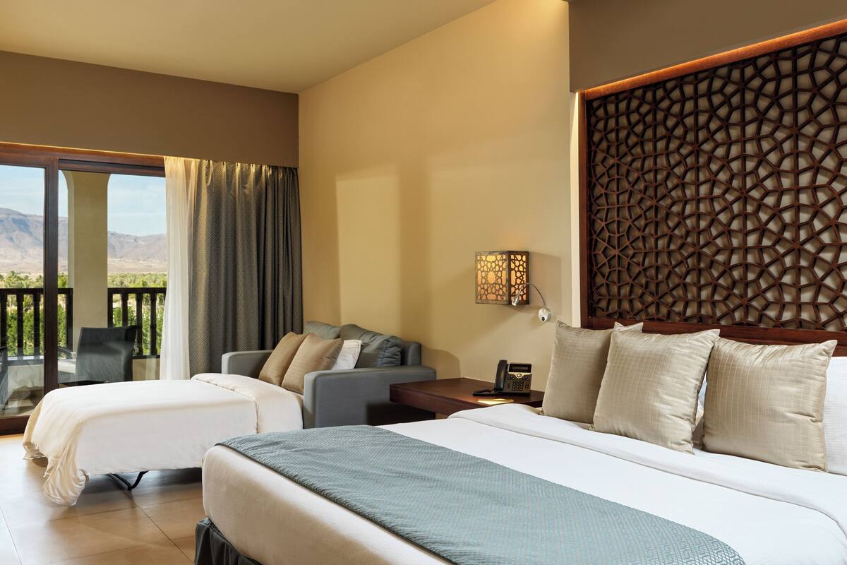 A hotel room with a large bed, a sofa, a desk, and a balcony with a view of the mountains.