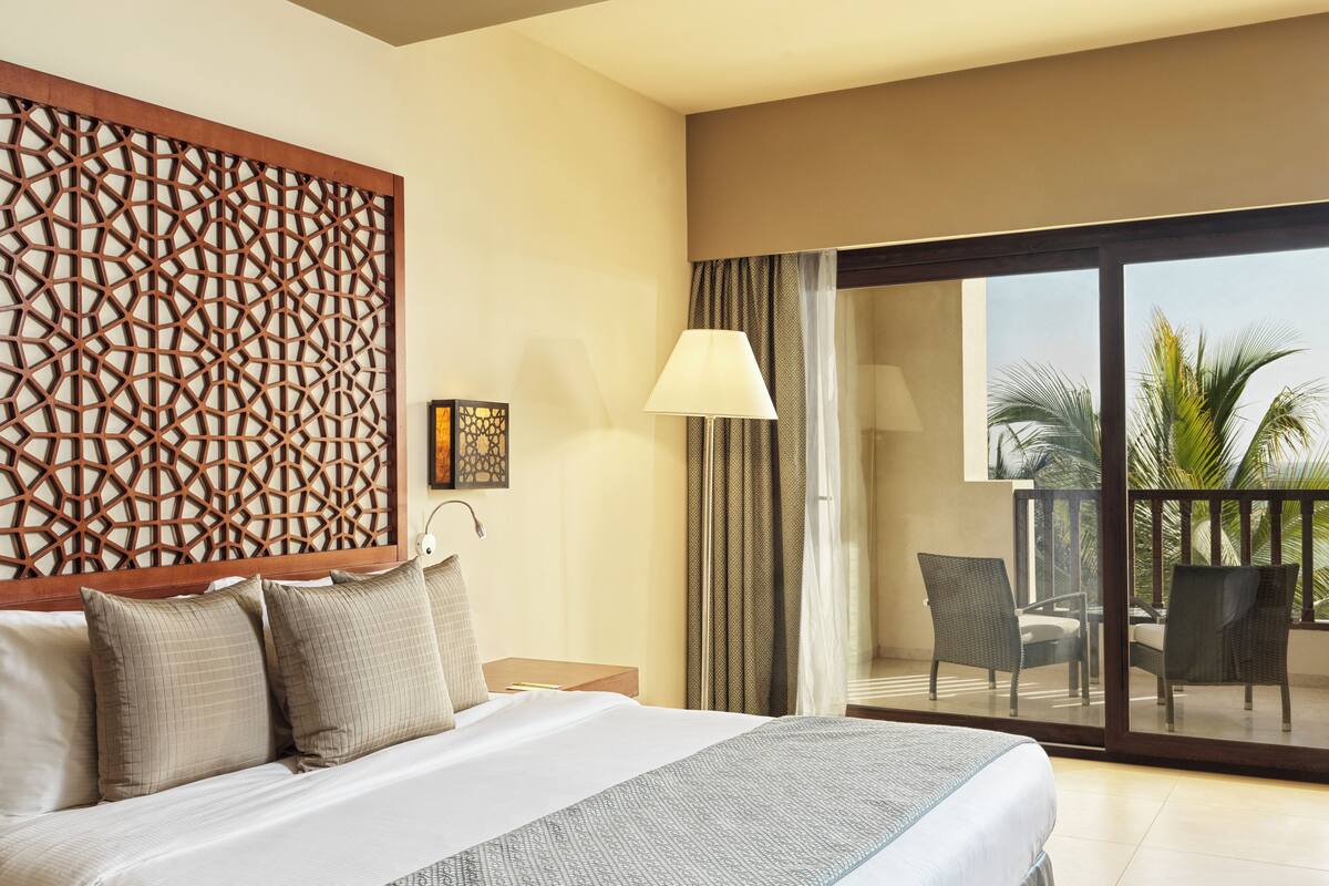 A hotel room with a bed, a nightstand, a lamp, a decorative wall panel, a sliding glass door leading to a balcony with chairs and a palm tree.
