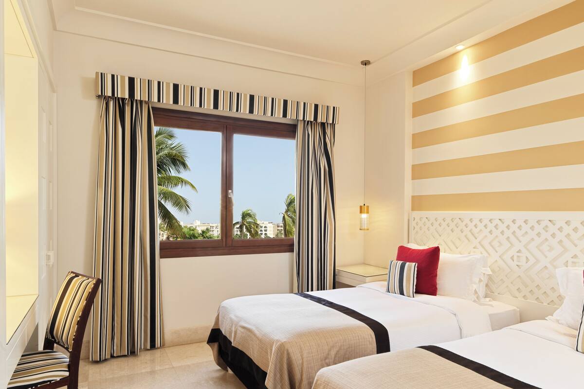 A hotel room with two beds, a chair, a window with striped curtains, a wall-mounted light, and a view of palm trees.