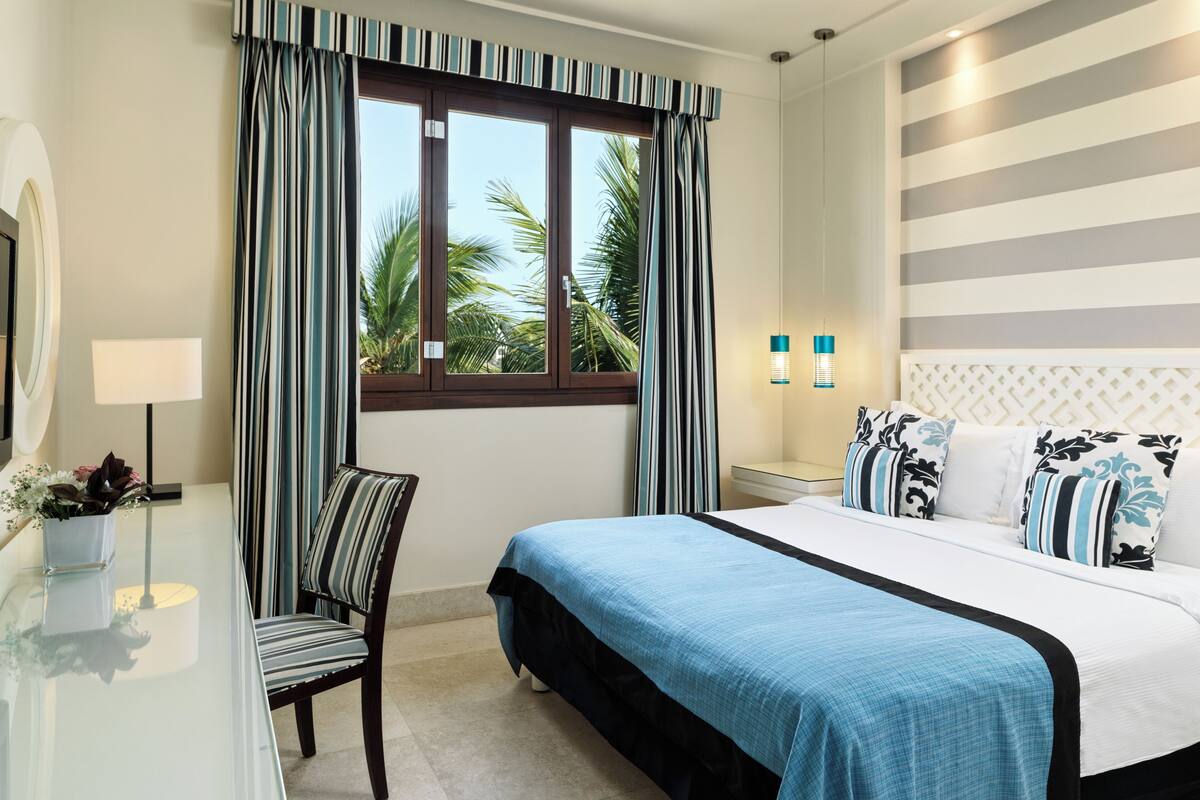 A hotel room with a bed, a chair, a desk with a lamp, and a window with curtains.