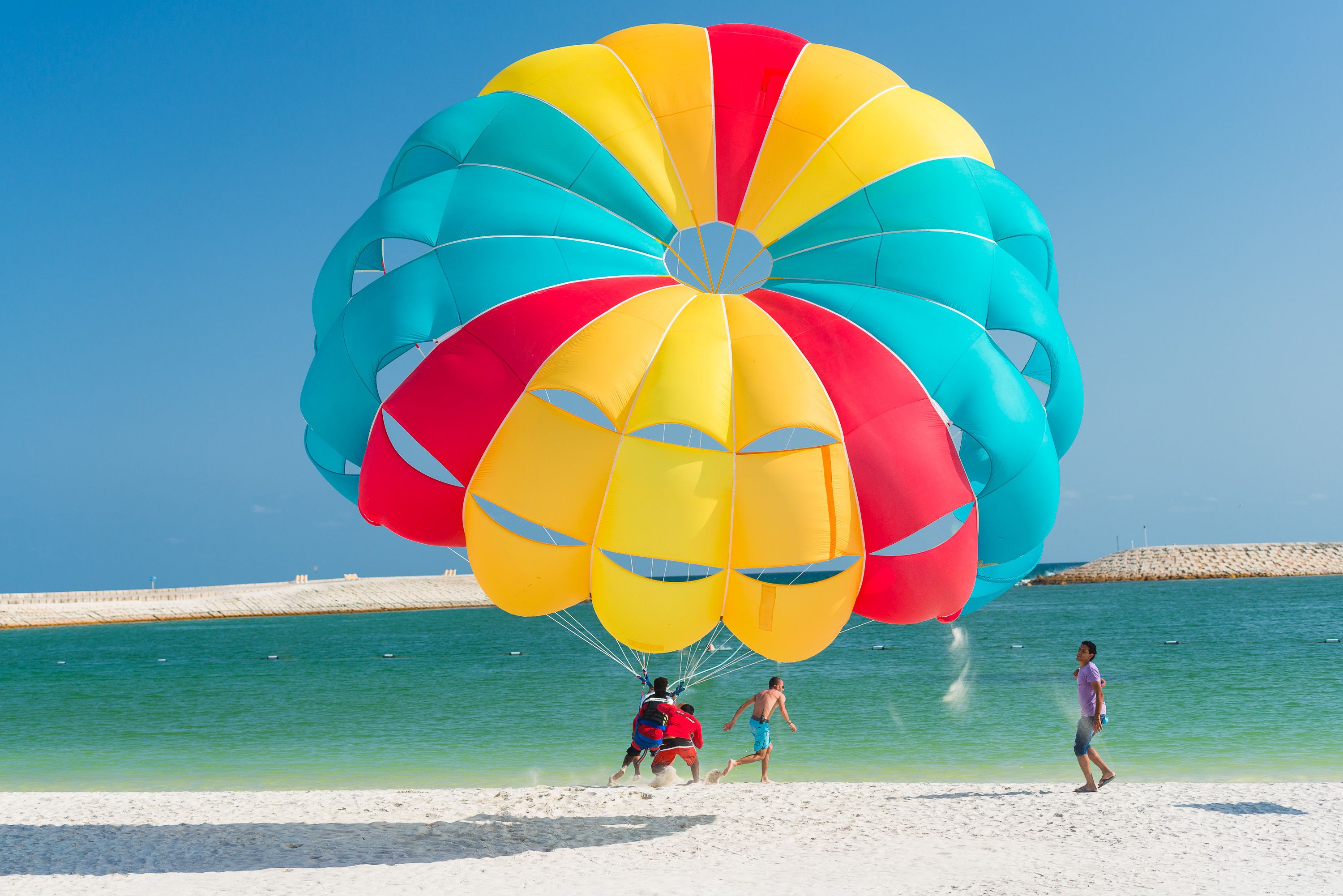 Une scène de plage avec une voile colorée, une eau d’un bleu limpide et une plage de sable.