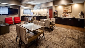 A hotel room with a dining area featuring patterned chairs and tables, a kitchen area with a coffee machine, and a television mounted on the wall. - Best Western Plus Stevens County Inn (Hugoton)