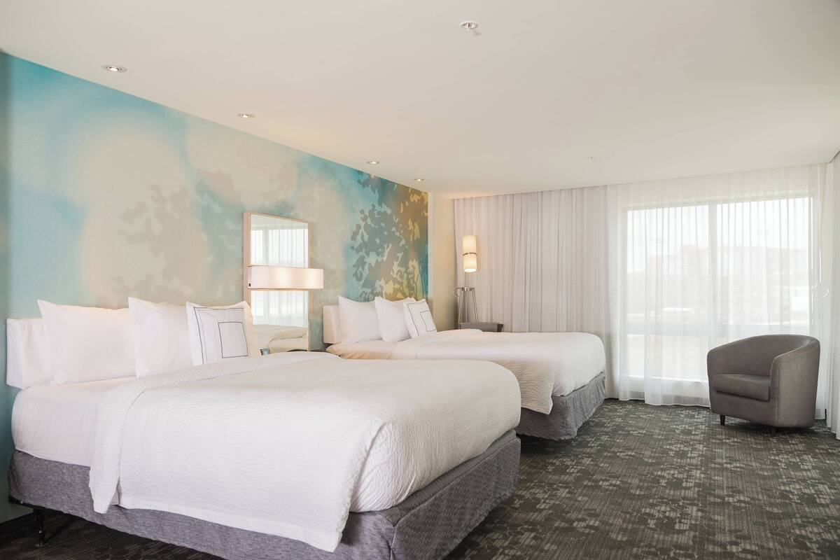 A hotel room with two beds, a chair, a large window with sheer curtains, and a wall with a blue abstract design.