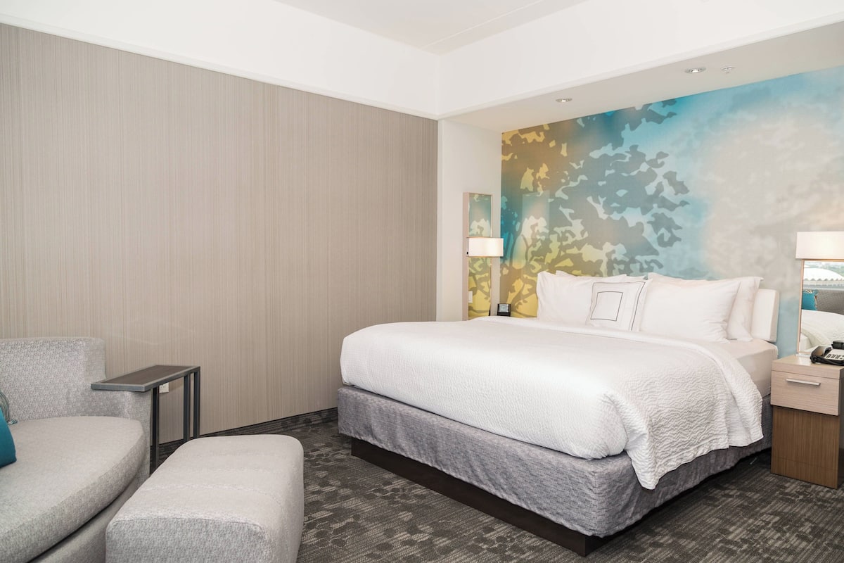 A hotel room with a large bed, a bedside table with a lamp, a chair, and a small table.