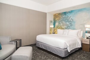A hotel room with a large bed, a bedside table with a lamp, a chair, and a small table.