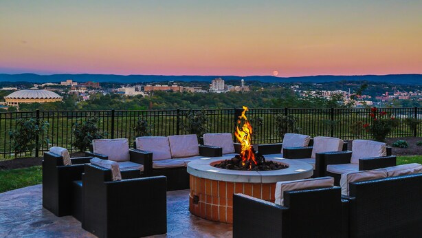 A patio area with outdoor seating and a central fire feature, overlooking a cityscape during sunset.