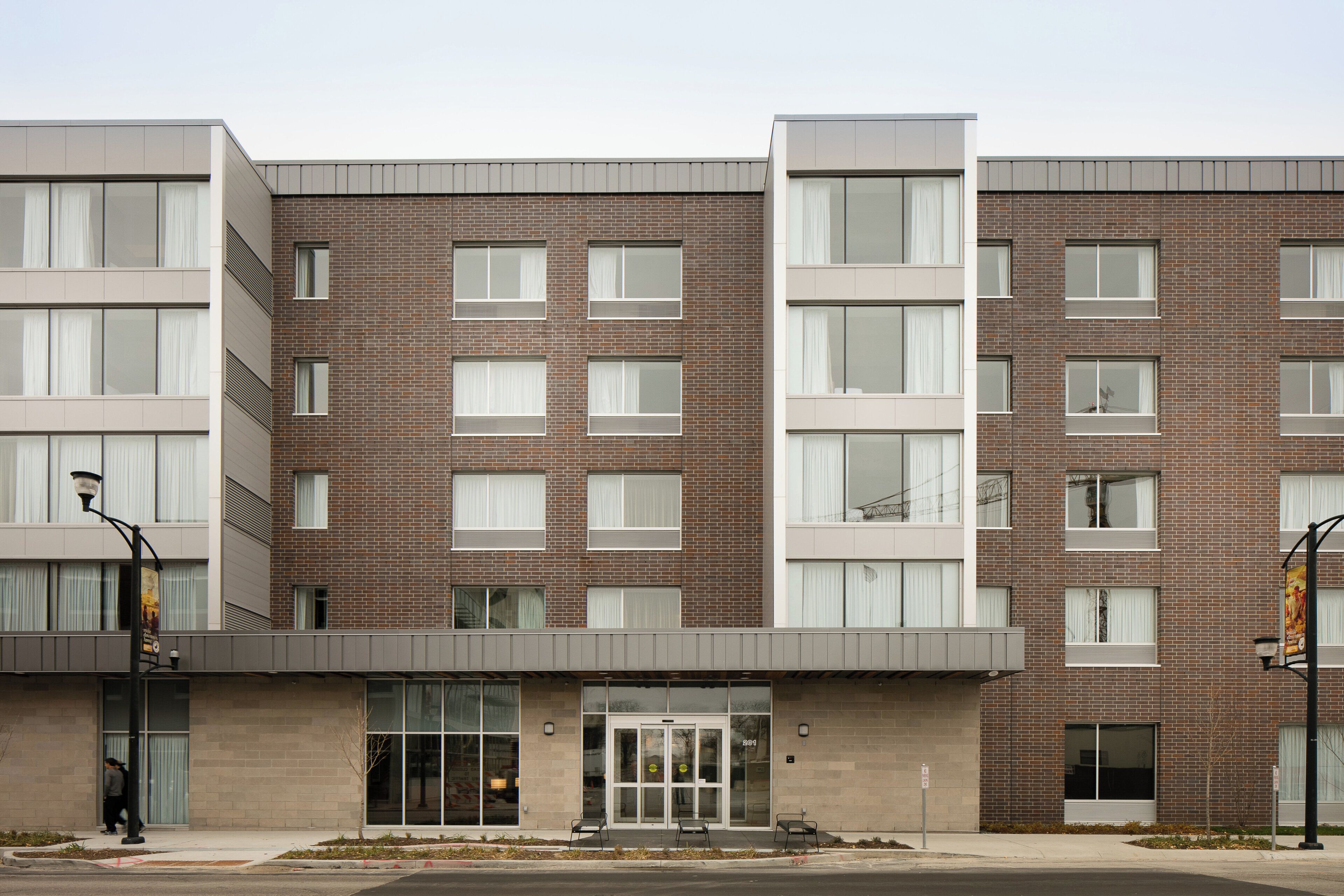 A multi-story building with a brick facade, large windows, and a flat entrance area with a door and two chairs outside.