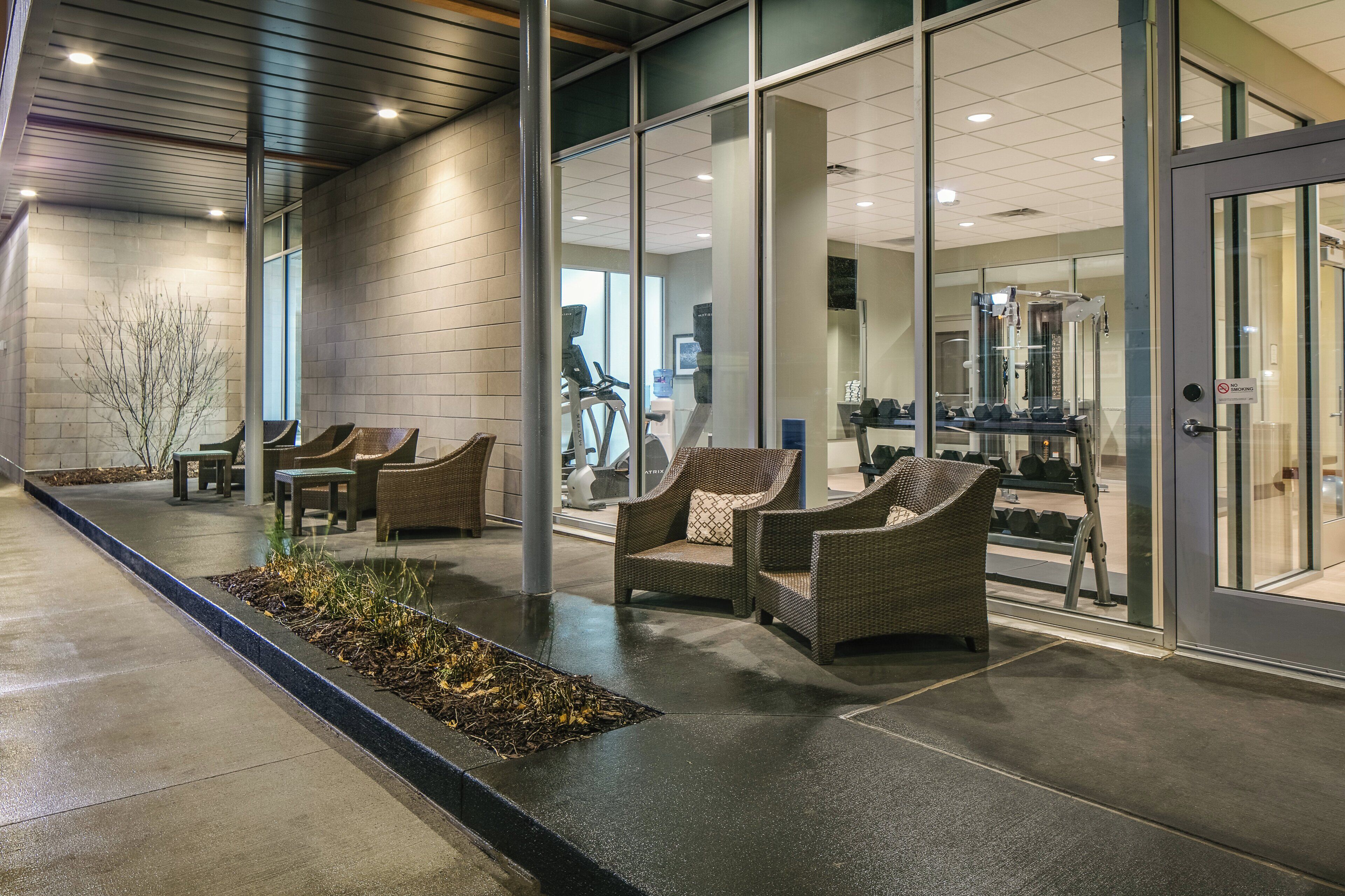 An indoor waiting area with wicker chairs, a concrete floor, and glass doors leading to a fitness area.