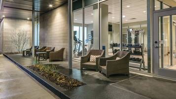 An indoor waiting area with wicker chairs, a concrete floor, and glass doors leading to a fitness area.