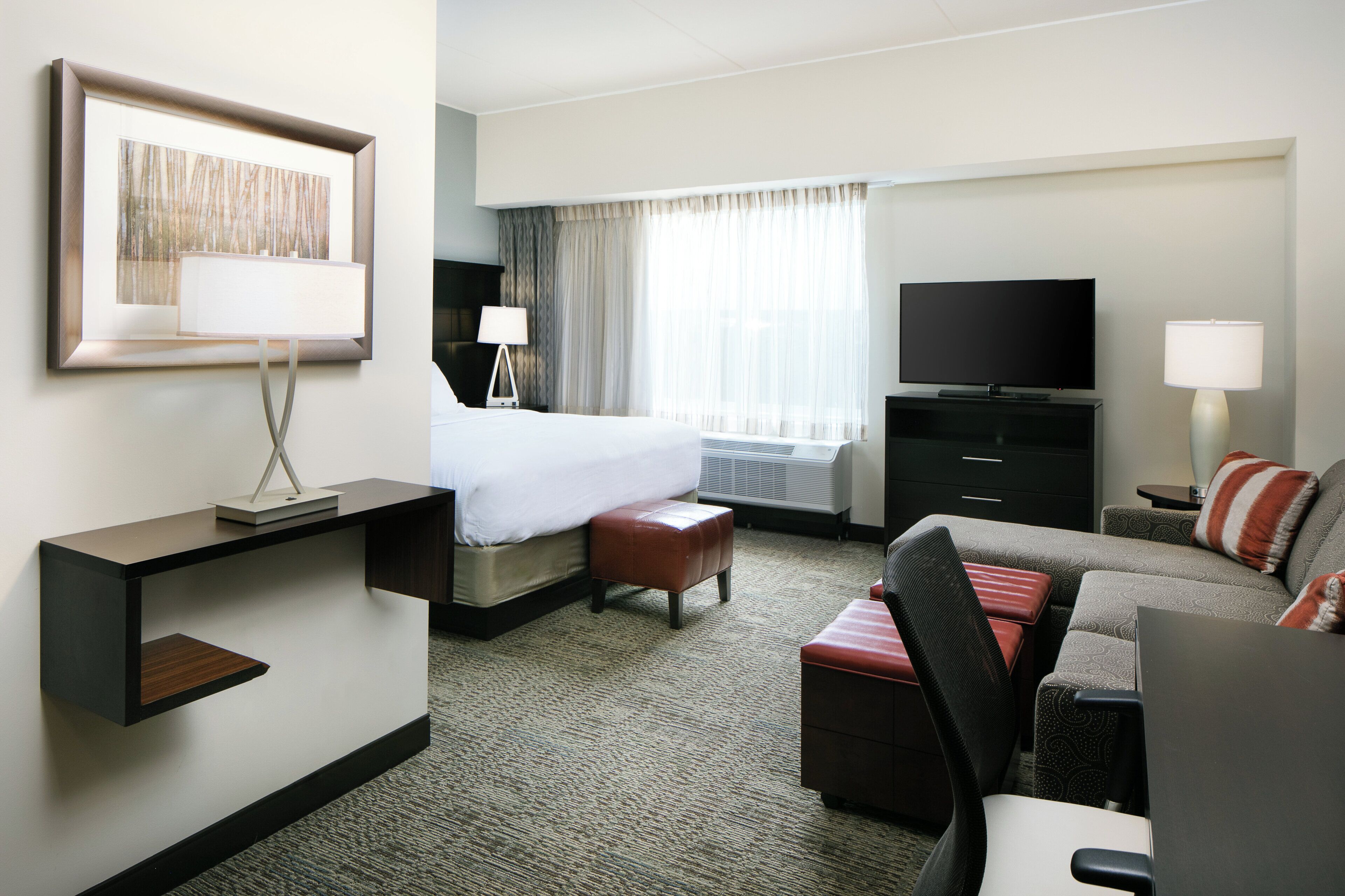 Quarto de hotel com cama, escrivaninha, televisão, sofá e uma janela grande com cortinas.