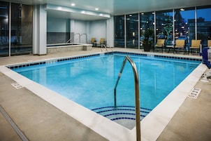 An indoor swimming pool with clear water, a metal ladder, and a view of the city at night through large windows.