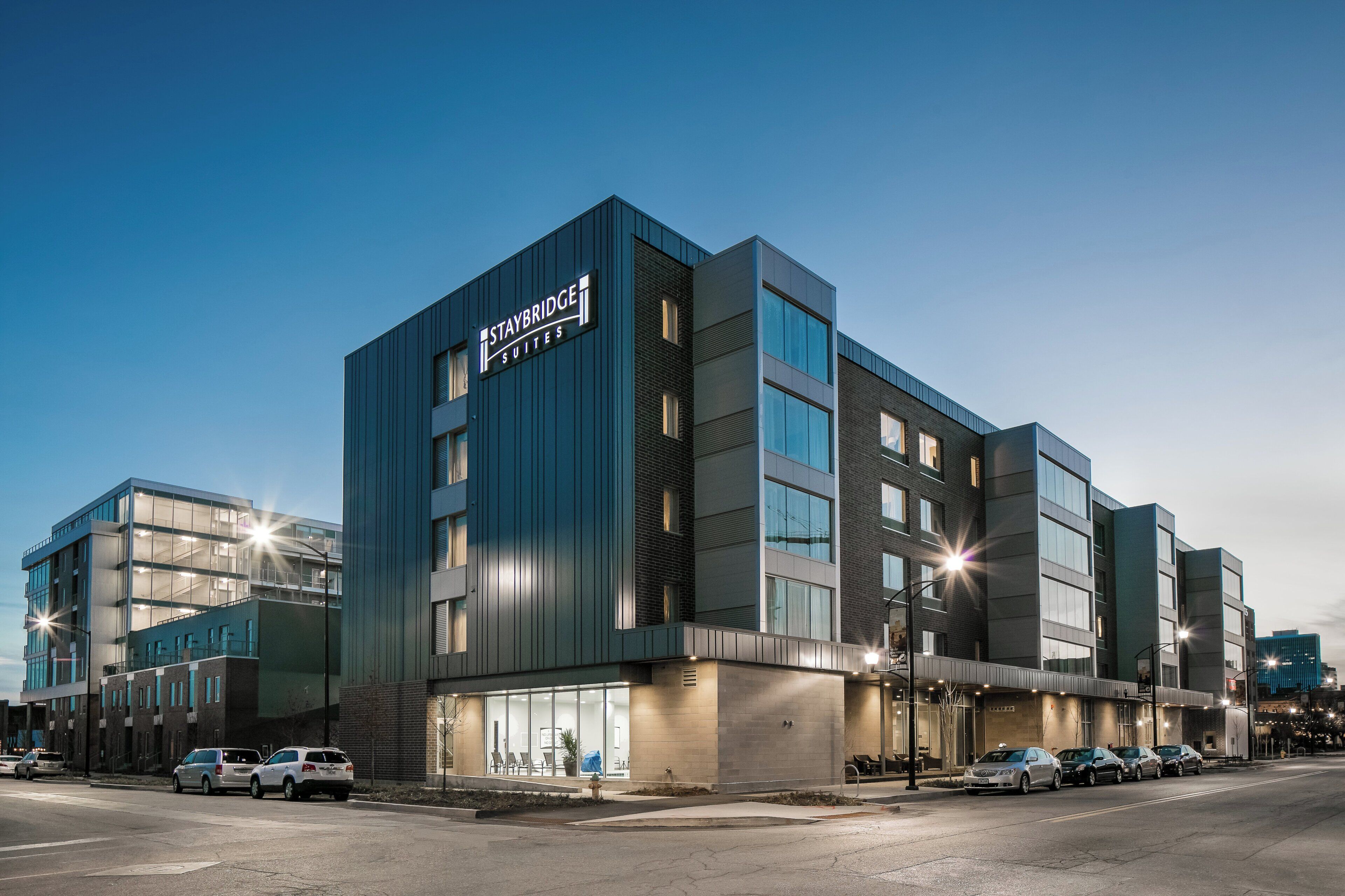 Um edifício moderno com uma placa que diz 'Staybridge Suites'. Apresenta uma combinação de fachada de vidro e tijolo, janelas amplas e um telhado plano.