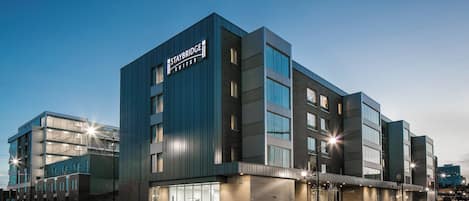 Un edificio moderno con un letrero que dice 'Staybridge Suites'. Presenta una combinación de fachadas de vidrio y ladrillo, ventanas grandes y un techo plano.