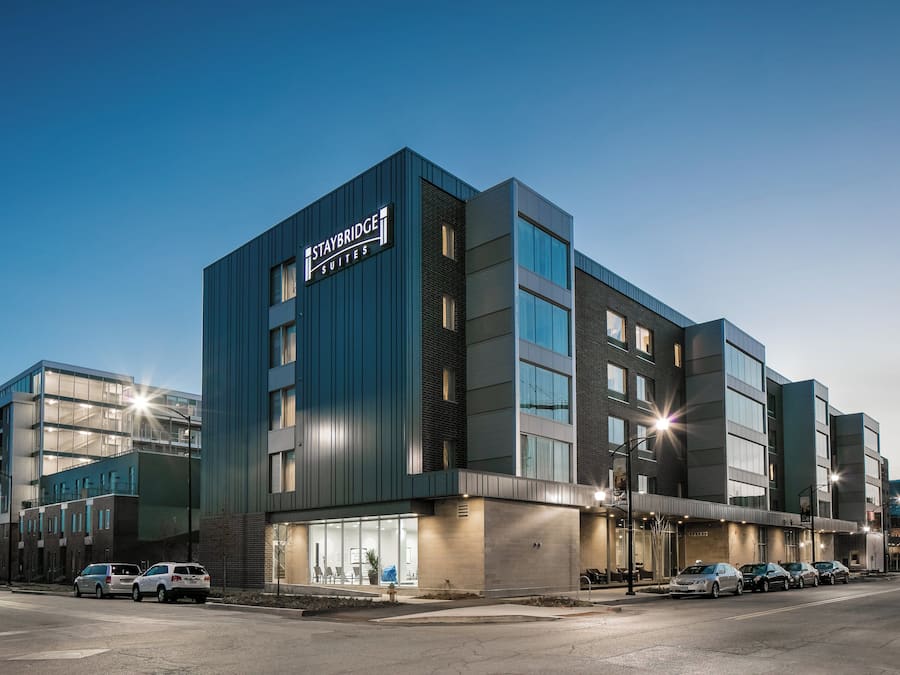 A modern building with a sign that reads 'Staybridge Suites.' It features a mix of glass and brick facades, large windows, and a flat roof.