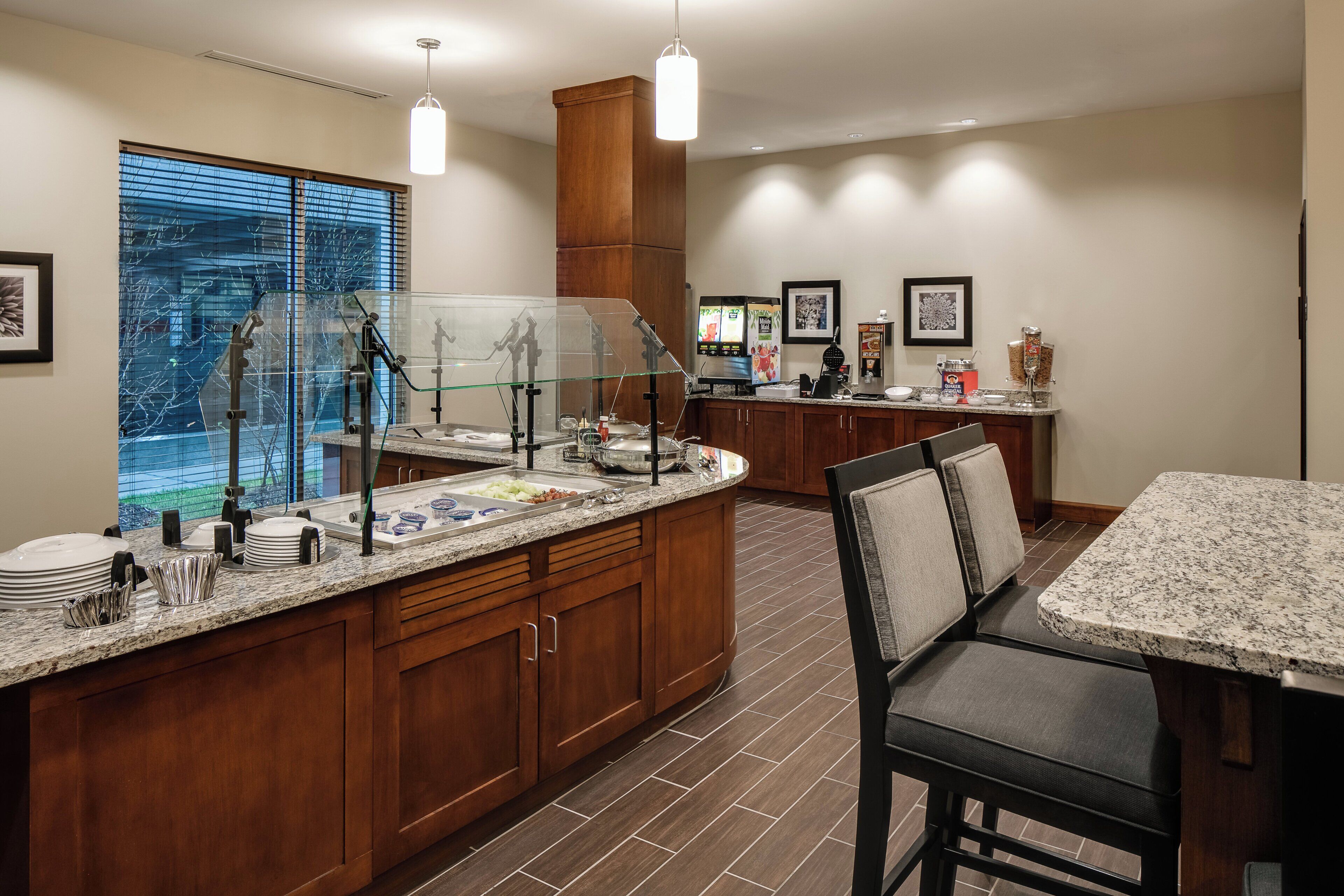 A hotel kitchen with a buffet setup, including a glass display case, a coffee station, and a seating area with a table and chairs.