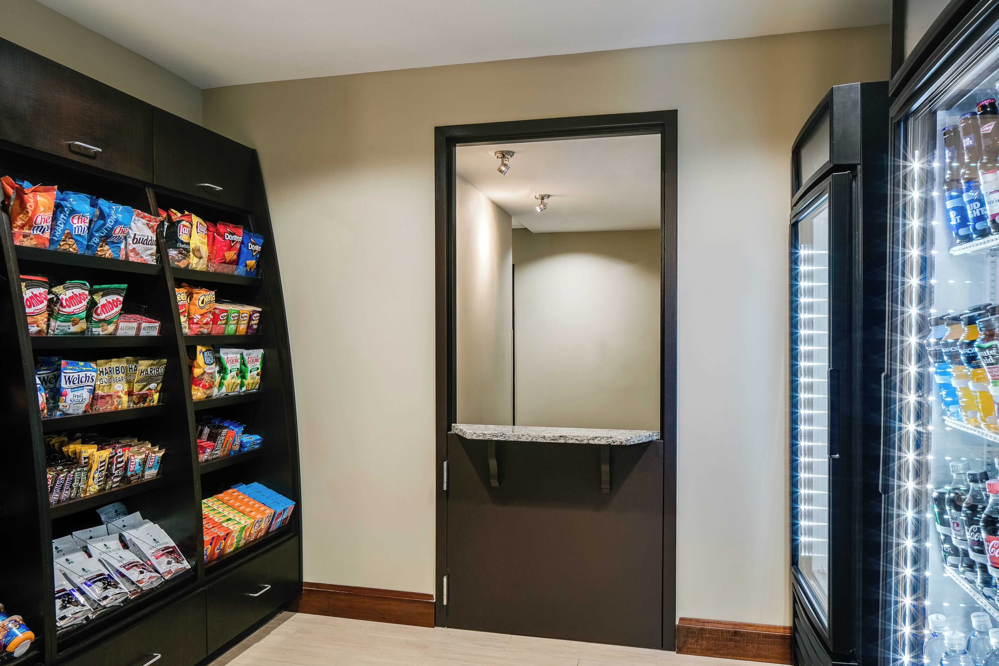 Um corredor com uma porta, prateleiras repletas de lanches e uma geladeira com bebidas.
