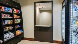 A hallway with a door, shelves stocked with snacks, and a refrigerator with beverages. - Staybridge Suites Des Moines Downtown by IHG (Des Moines)