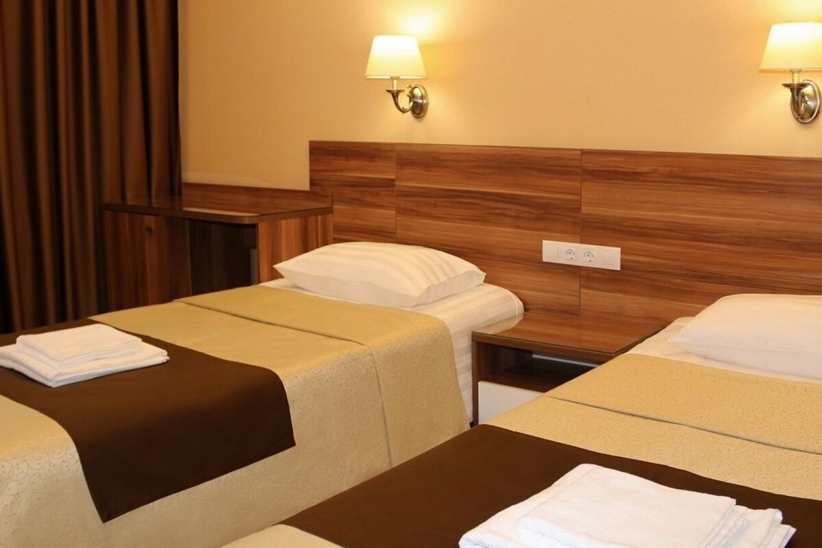 A hotel room with two beds, each with a brown headboard, white linens, and a small bedside table. There are wall-mounted lamps on either side of the headboard.