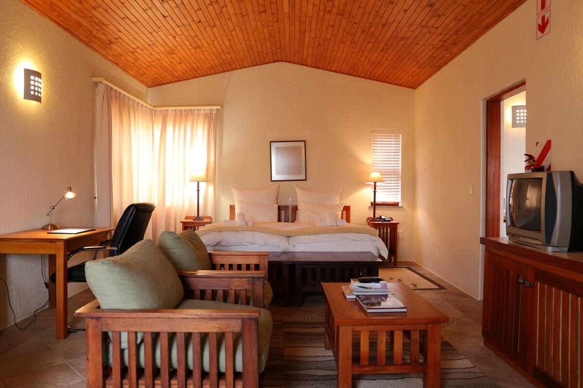 Habitación de hotel con techo de madera, cama con ropa de cama blanca, escritorio con lámpara, televisor sobre un mueble de madera y una silla.