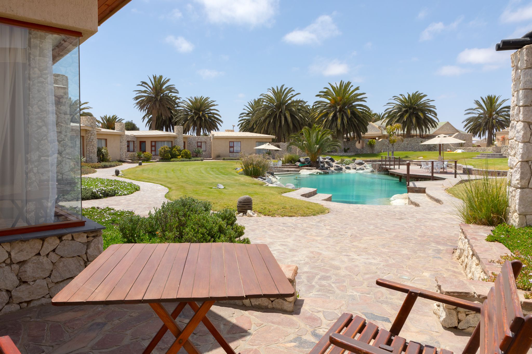A pool area with stone pathways, wooden furniture, and palm trees.