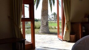Un dormitorio con cama, televisión y vistas a palmeras y campos a través de cortinas abiertas.