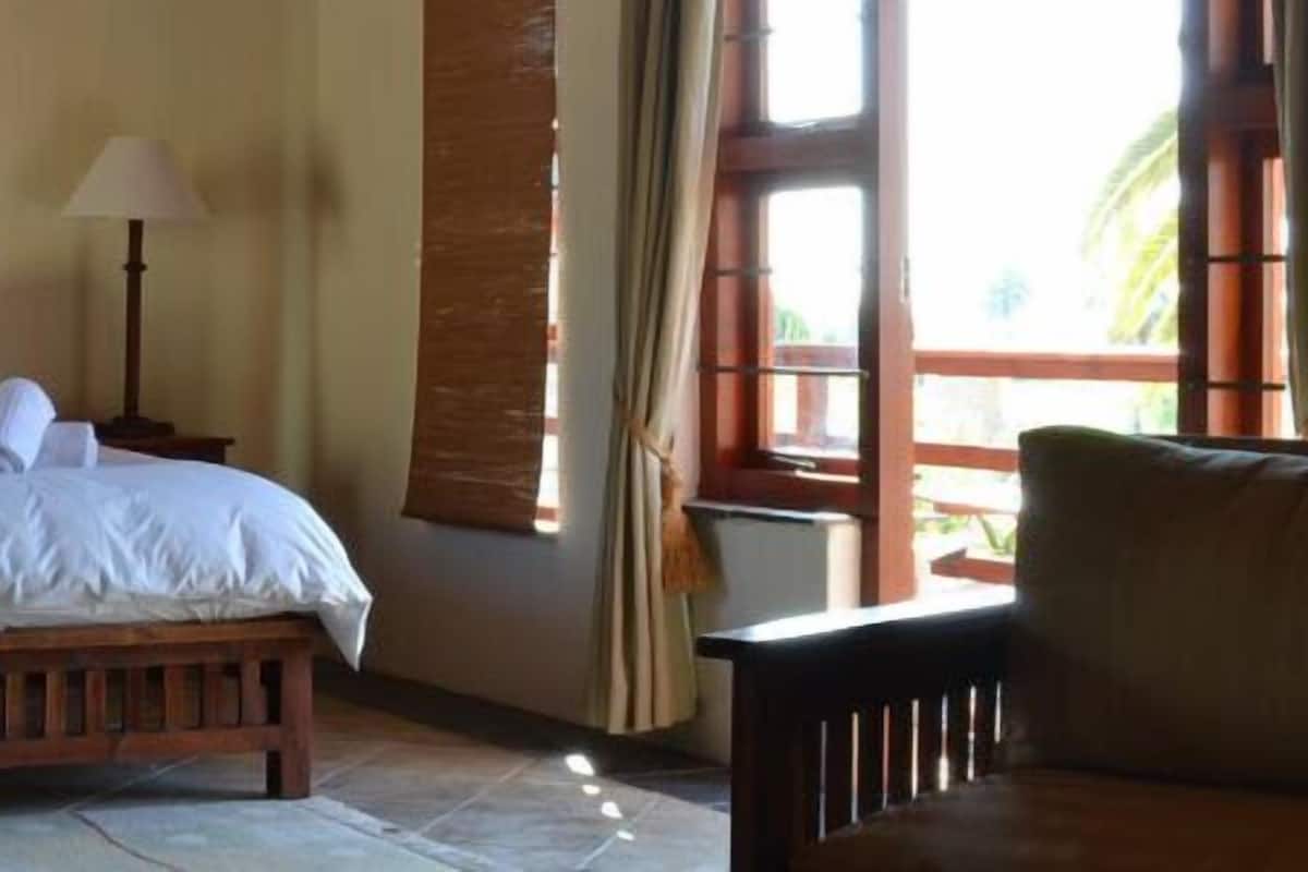 Una cama doble con sábanas blancas, dos mesitas de noche con lámparas, un banco de madera y vistas al exterior a través de una ventana.