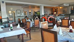 A restaurant interior with wooden chairs and tables, a clock on the wall, and life rings on the back wall. - The Chapman Hotel & Conference Centre (Gqeberha (Port Elizabeth))